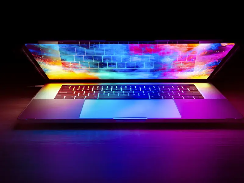 An open laptop on a dark desk, its screen and keyboard glowing with a colourful design